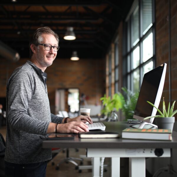 Homme en pull gris travaillant debout devant un ordinateur à un bureau réglable dans un open space industriel lumineux