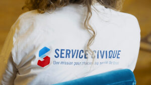 Dos d'une jeune femme blonde portant un t-shirt blanc avec le logo Service Civique et le slogan « Une mission pour chacun au service de tous »
