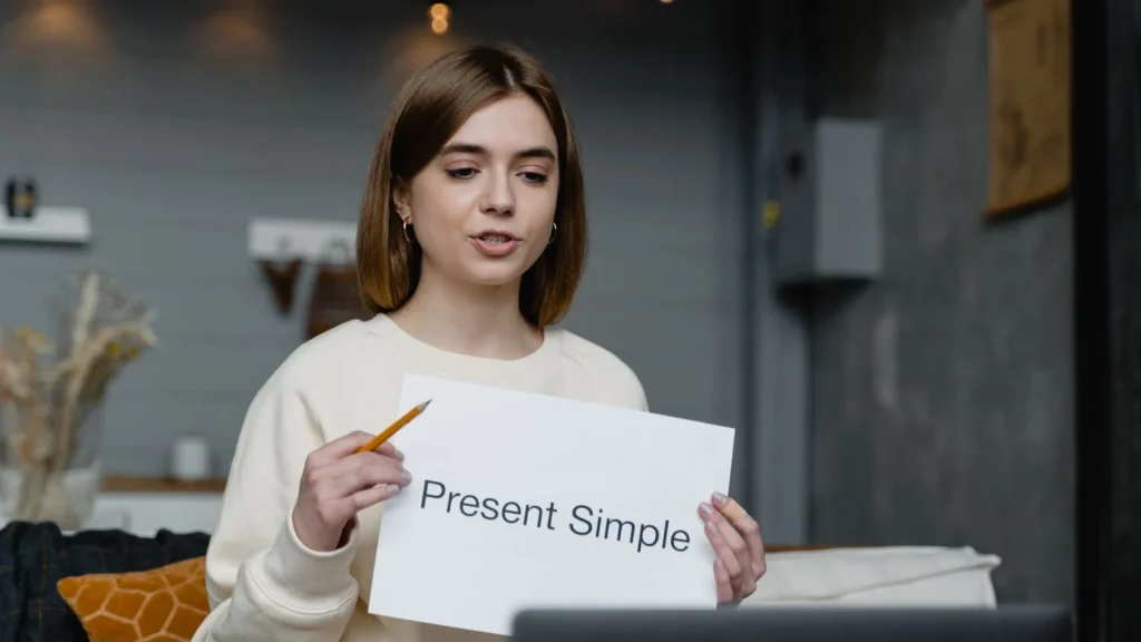 Une enseignante montre une fiche « Present Simple » devant un ordinateur portable.