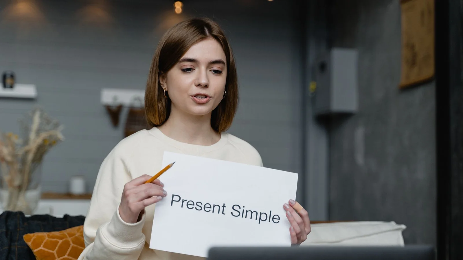 Une enseignante montre une fiche « Present Simple » devant un ordinateur portable.