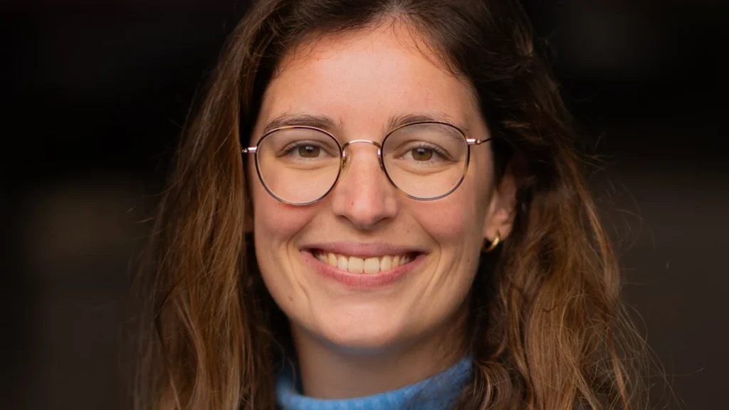 Portrait de Clotilde Mérillon, souriante, avec des lunettes et un pull bleu.