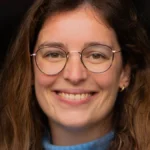 Portrait de Clotilde Mérillon, souriante, avec des lunettes et un pull bleu.