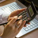 Mains utilisant une calculatrice noire devant un ordinateur portable, avec un stylo entre les doigts.
