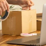 Préparation d’un colis devant un ordinateur, illustrant les frais de traitement des colis importés.