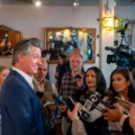 Gavin Newsom parle devant des microphones, entouré de plusieurs personnes lors d’une conférence de presse.