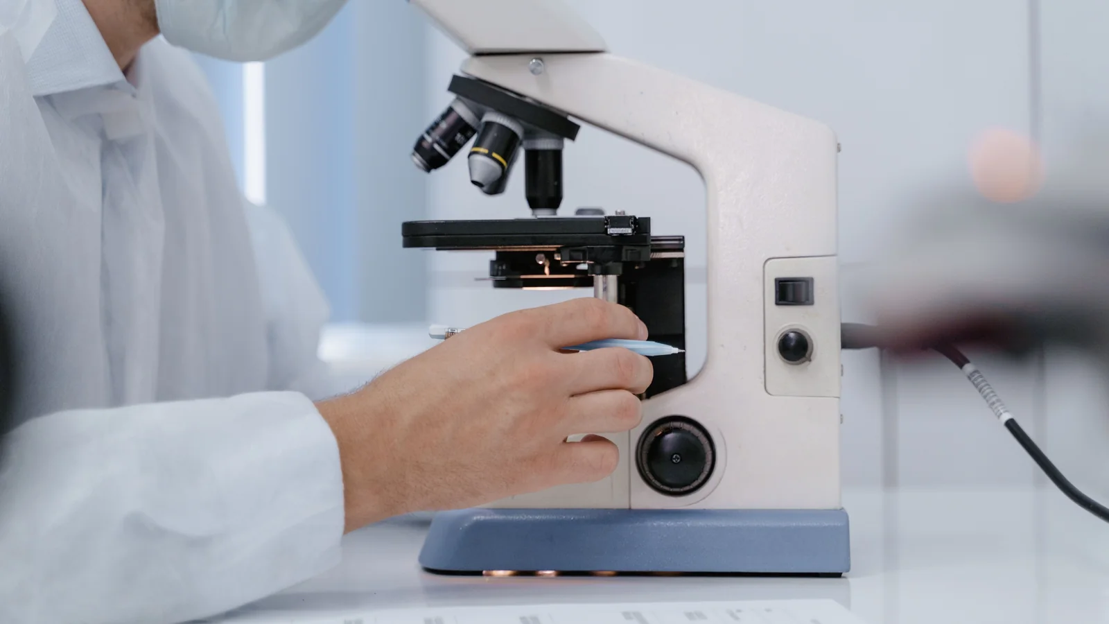 Une personne en blouse et masque manipule un microscope en laboratoire.