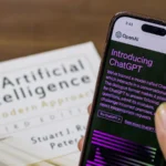 Une main tient un smartphone affichant la présentation de ChatGPT, devant un livre sur l’intelligence artificielle.