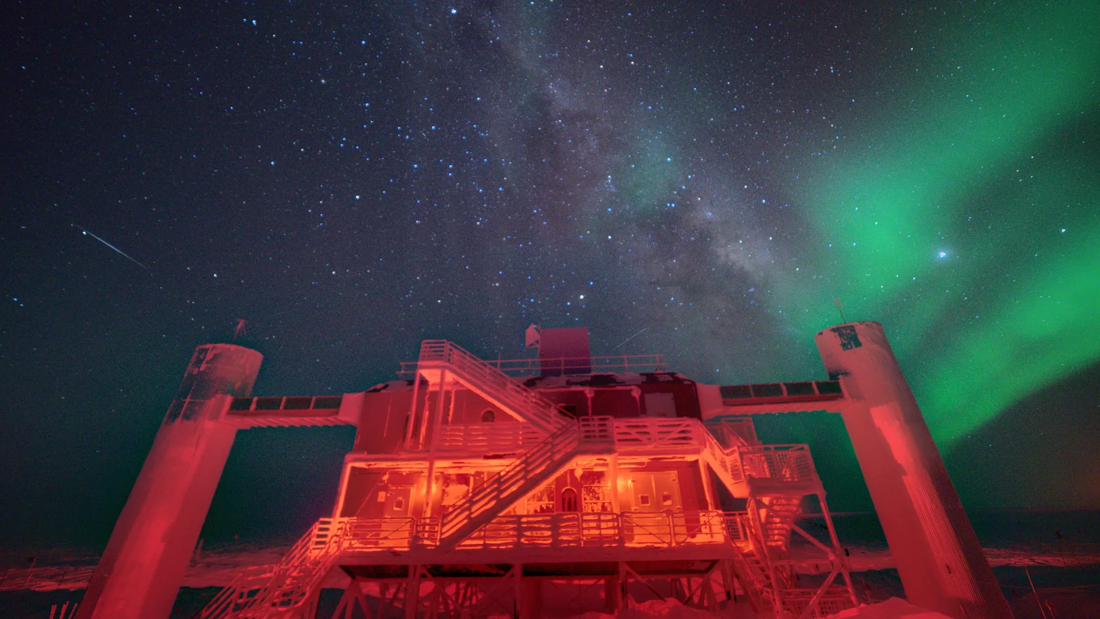 Le laboratoire IceCube éclairé en rouge au pôle Sud, sous la Voie lactée et une aurore australe.