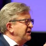 Jean-Luc Mélenchon de profil, lunettes et cravate rouge, lors d’une réunion publique.