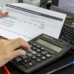 Une personne utilise une calculatrice devant des documents financiers et un écran affichant des graphiques.