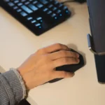 Une main posée sur une souris noire, à côté d’un clavier et d’un ordinateur portable.