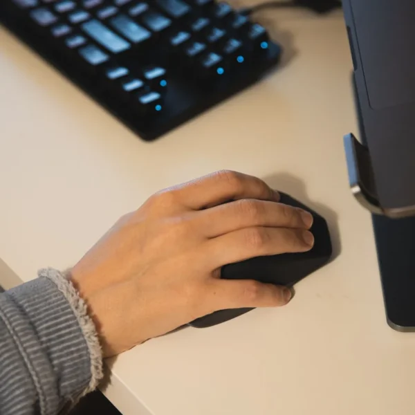 Une main posée sur une souris noire, à côté d’un clavier et d’un ordinateur portable.