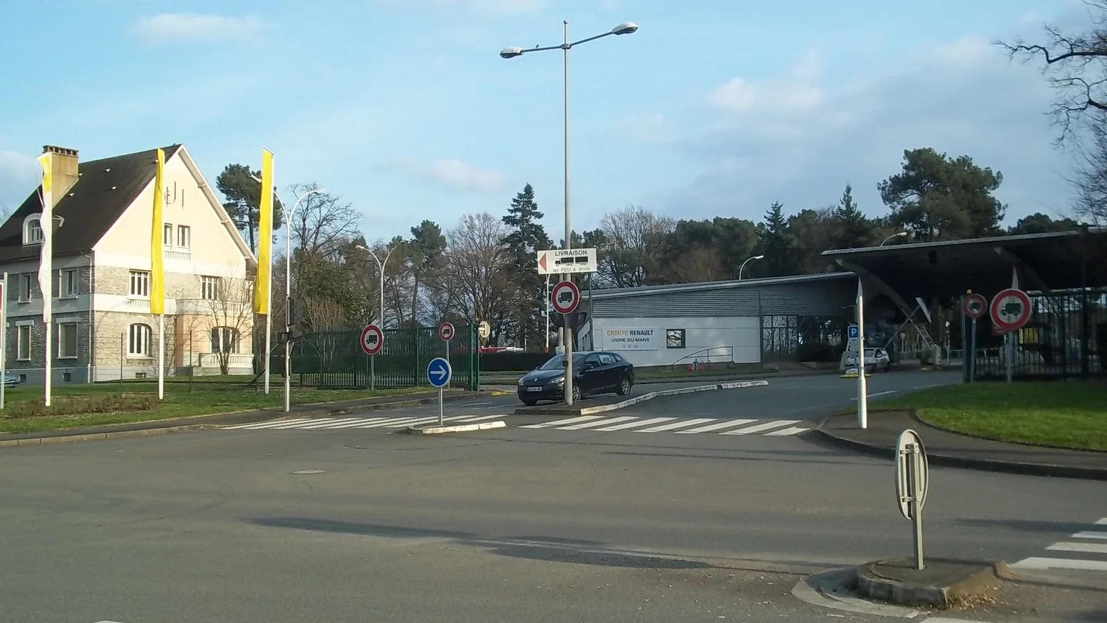 Entrée de l’usine Renault du Mans, avec ses bâtiments, son portail et des drapeaux jaunes.