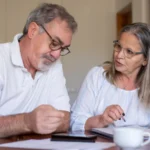 Un couple de seniors consulte des documents à une table, avec un téléphone posé devant lui.