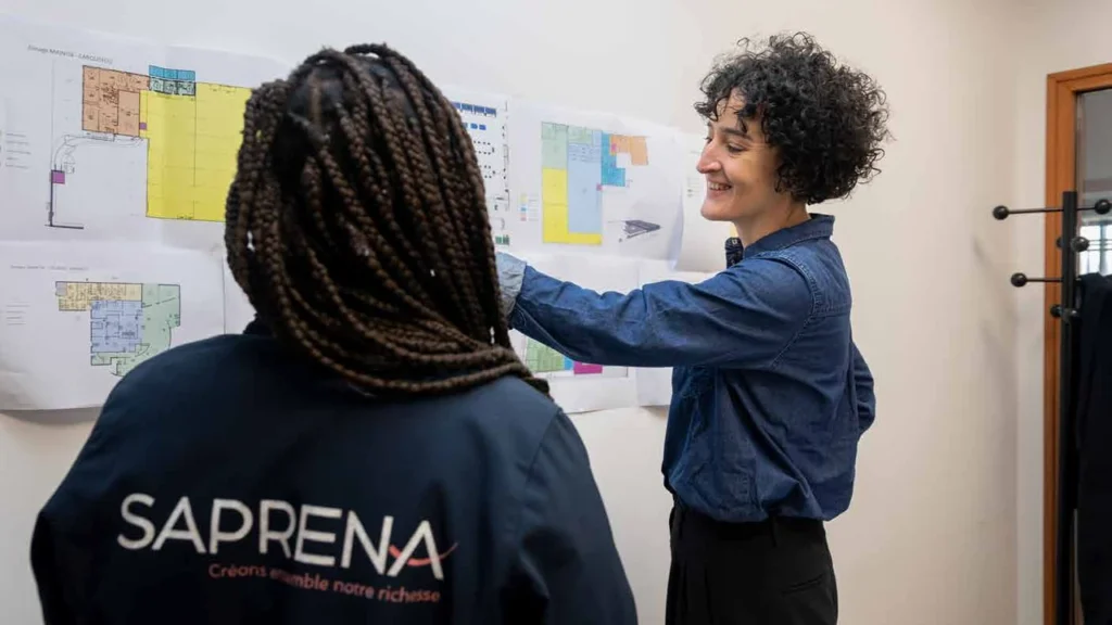 Deux personnes échangent devant des plans au mur, l’une portant une veste avec le logo SAPRENA.
